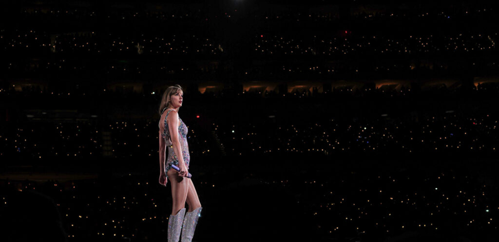 Taylor Swift de cuerpo completo sobre el escenario durante The Eras Tour, con el publico iluminando el estadio detras